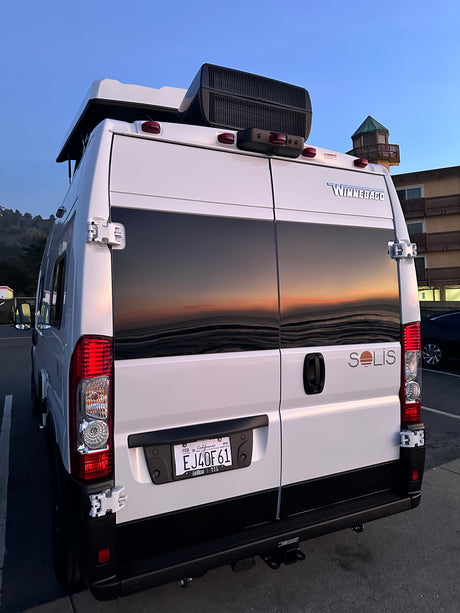 White Winnebago Solis camper van for **rv rental**, its rear windows reflecting a stunning sunset over water.