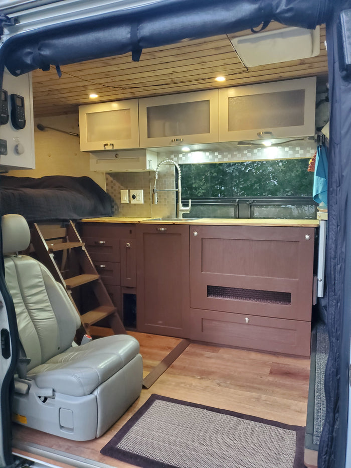 RV rental interior: Elevated bed with ladder, full kitchenette, and a comfortable swiveling seat in a cozy camper van.