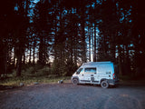 An RV rental camper van parked on a gravel path in a tall forest at sunset, perfect for outdoor travel.