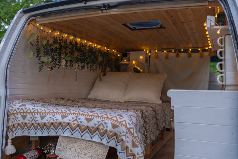 View of a cozy rv rental's rear interior: patterned bed with pillows, string lights, decorative plants, and custom wood ceiling. — photo 13
