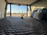 RV rental: Relax in a comfy bed in your camper van, enjoying desert views with saguaro cacti. Laptop on bed for convenience.