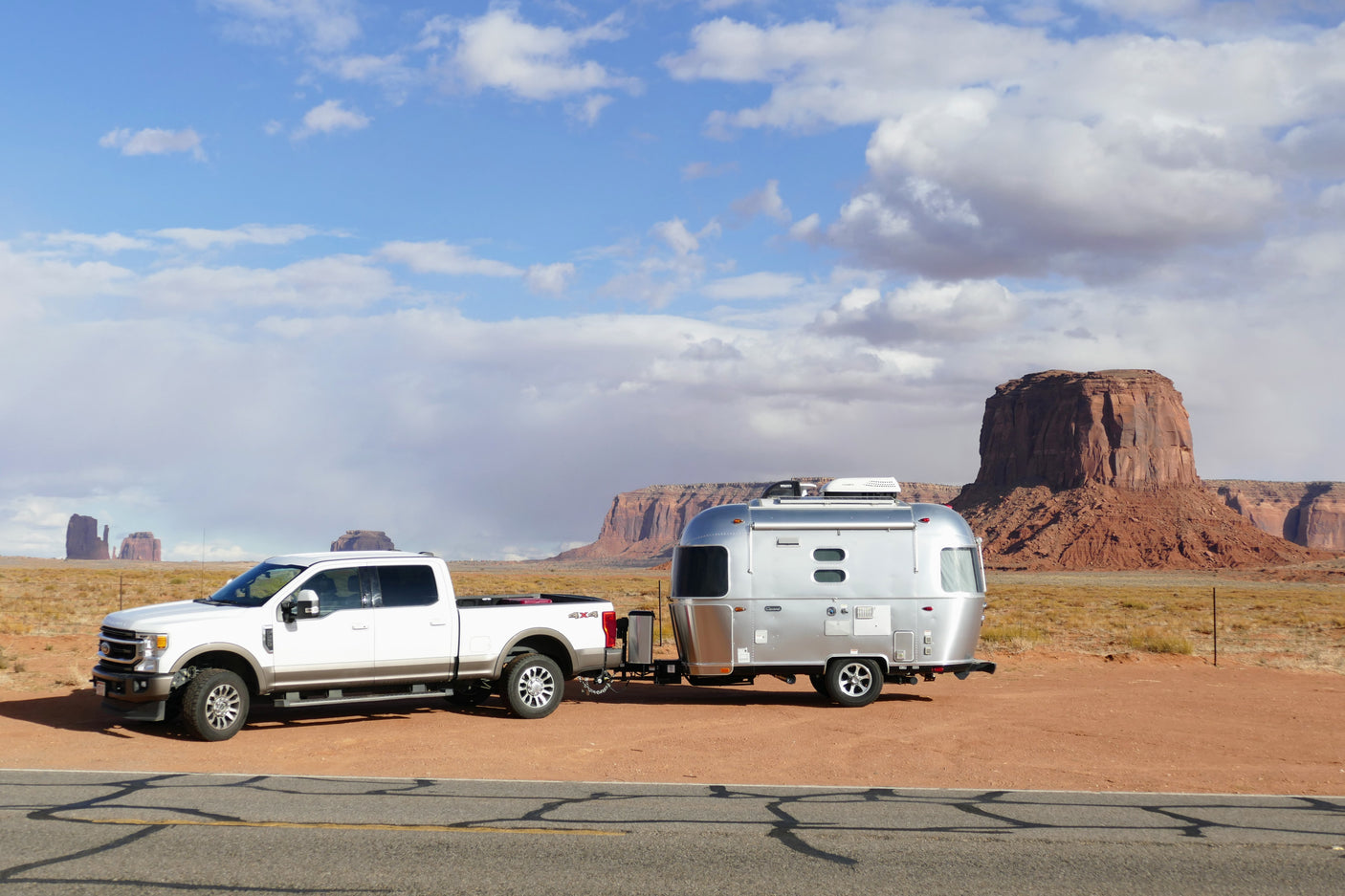 RV rental adventure in Monument Valley! A white truck tows a silver Airstream travel trailer against red rock formations under a cloudy sky.