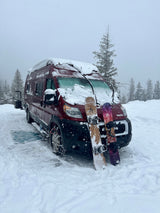 Red RV rental camper van blanketed in snow with two snowboards leaning, ready for adventure in a snowy forest.