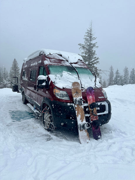 Red RV rental camper van blanketed in snow with two snowboards leaning, ready for adventure in a snowy forest.