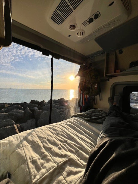 Golden hour bliss! Relax in your RV rental. Inside a van, person's legs stretch on a comfy bed overlooking a sunset ocean.