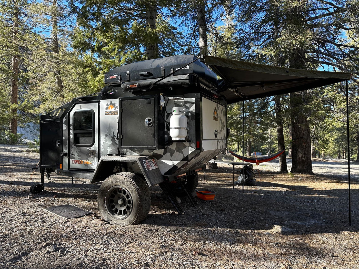 Rugged off-road camper trailer (RV rental) with camo design, rooftop tent, and awning at a sunny wooded campsite. — photo 12