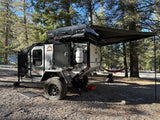 Rugged off-road camper trailer (RV rental) with camo design, rooftop tent, and awning at a sunny wooded campsite.