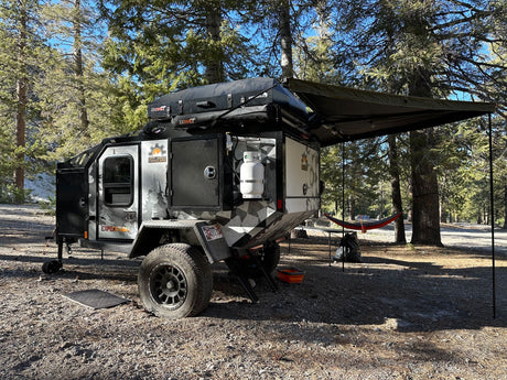 Rugged off-road camper trailer (RV rental) with camo design, rooftop tent, and awning at a sunny wooded campsite.