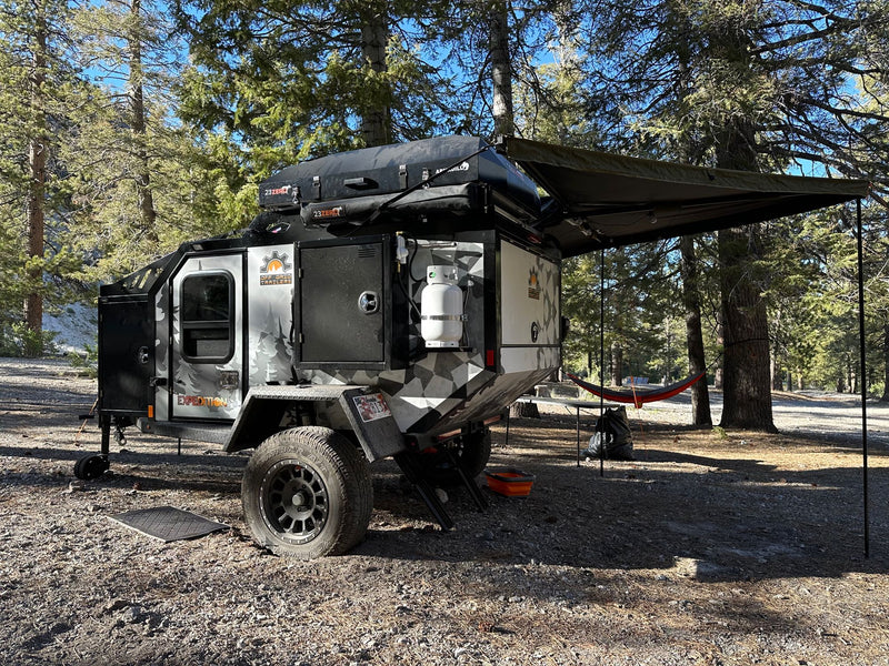 Rugged off-road camper trailer (RV rental) with camo design, rooftop tent, and awning at a sunny wooded campsite. — photo 12