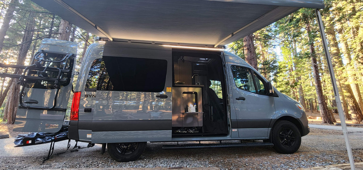 Grey camper van RV rental in a forest campsite with awning extended and interior visible. Ready for adventure!