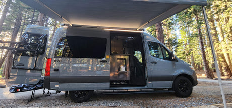Grey camper van RV rental in a forest campsite with awning extended and interior visible. Ready for adventure!