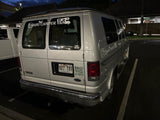 This Hawaiian rv rental is a silver Ford E250 camper van with "Oahu Camper Van" decals and QR codes on the back, parked at night.