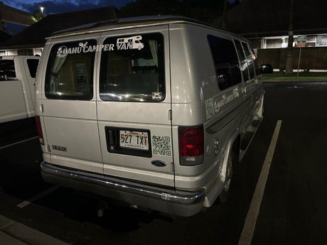 This Hawaiian rv rental is a silver Ford E250 camper van with "Oahu Camper Van" decals and QR codes on the back, parked at night.