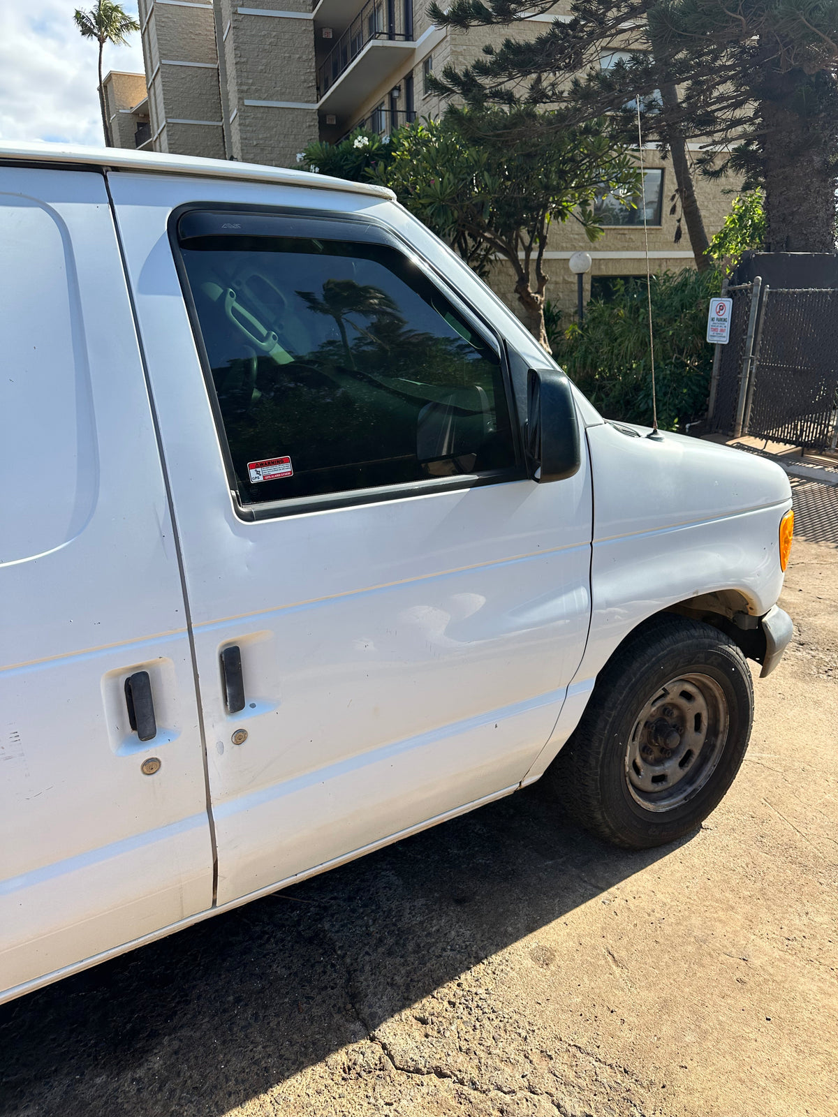White van (possible RV rental conversion) parked. Side view shows GPS alarm sticker, tire; building & trees in background. — photo 6