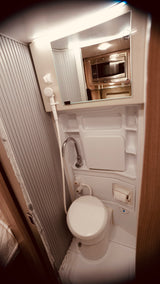 An RV rental's compact wet bath showing a toilet, shower, and sink. A mirror reflects a microwave.