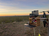 RV rental adventure: portable outdoor kitchen, camping stove, and bike set in a vast desert landscape under a beautiful sunset sky.