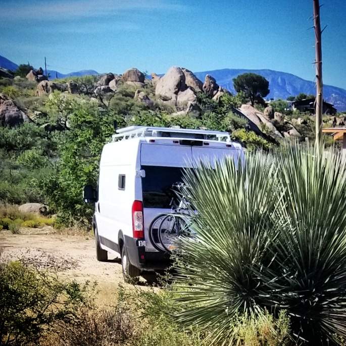 White RV rental van, with bikes, parked on a dirt track amidst arid rocky terrain & distant mountains. Ideal for adventure.