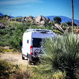 White RV rental van, with bikes, parked on a dirt track amidst arid rocky terrain & distant mountains. Ideal for adventure.