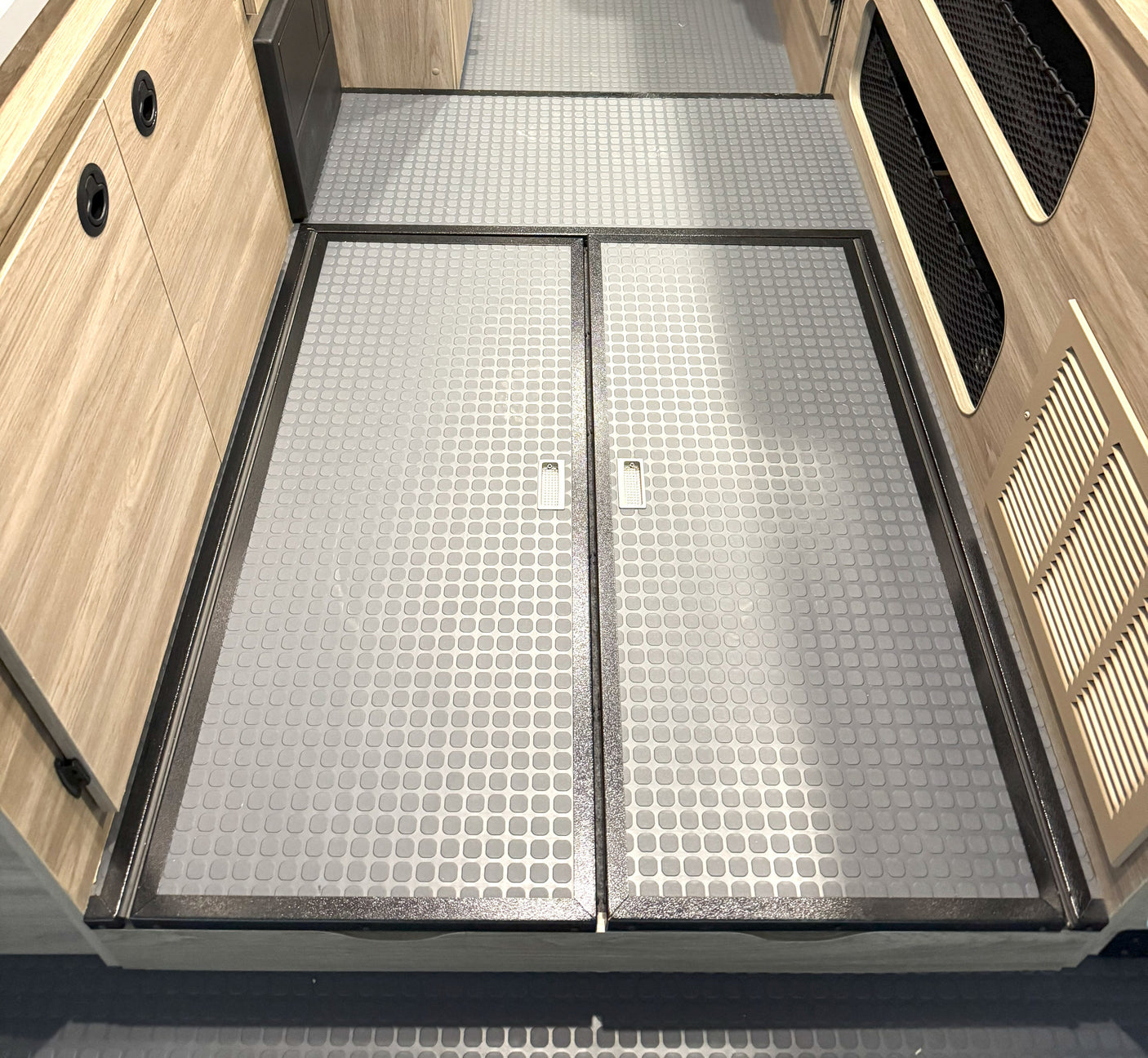 Detailed view of an RV rental's grey, non-slip floor with framed access hatches and pull tabs, flanked by wood paneling.
