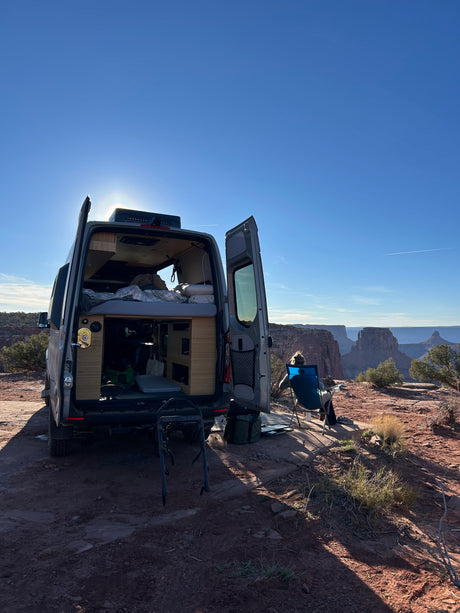 This RV rental provides a perfect view. A camper van is parked with open doors, revealing a cozy interior as a person enjoys the vast canyon vista.