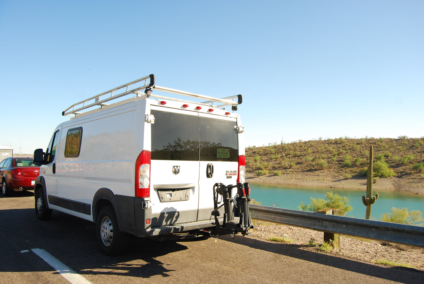 This RV rental (white van with bike rack & roof rack) is parked roadside by a desert lake in Arizona, under a clear sky.