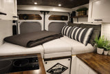 Cozy RV rental camper van interior. Features a comfy bed, modern kitchen with induction cooktop, and wood finishes.