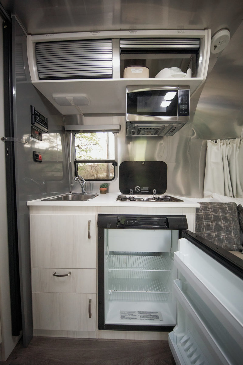 This rv rental's compact kitchen features a sink, stove, microwave, and open fridge with labeled overhead storage. — photo 11