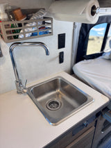 RV rental interior: Kitchenette with sink, faucet, paper towels, storage, and power outlets. Bed in background.