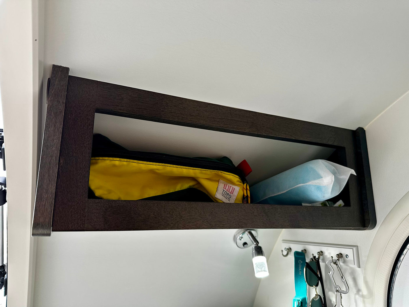 RV rental overhead storage cabinet contains a yellow pouch and light blue item. A reading light and hooks are below.