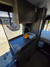 Compact RV rental interior featuring a kitchen sink, bed area, and an open side door view of an electric car outside.