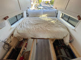 This RV rental features a cozy bed with clean white bedding, overhead cabinets, and essential under-bed storage for mobile living.