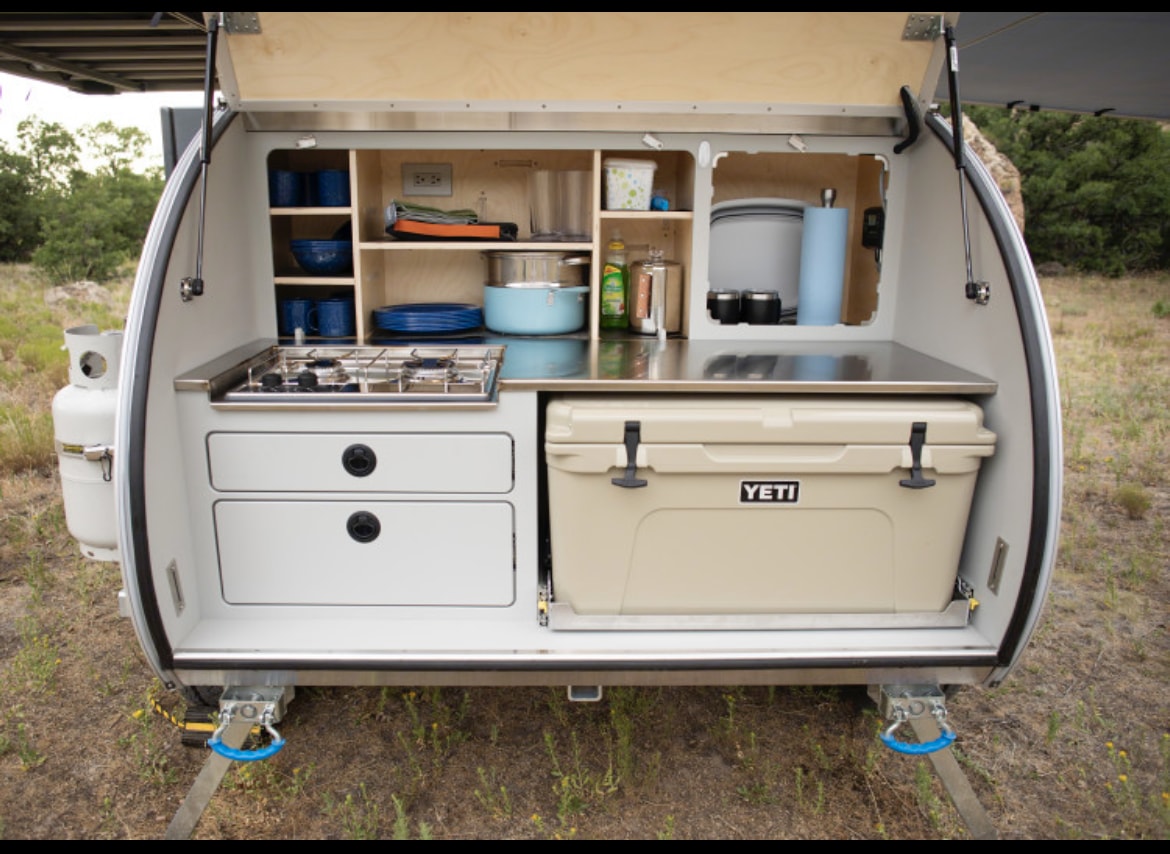Compact RV rental teardrop trailer shows its rear outdoor kitchen: stove, counter, shelves, and pull-out YETI cooler. Great for camping. — photo 4