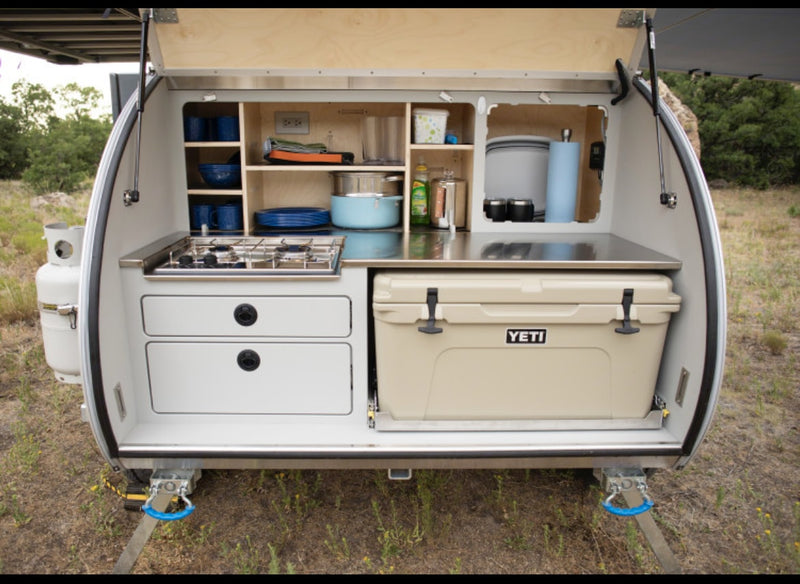 Compact RV rental teardrop trailer shows its rear outdoor kitchen: stove, counter, shelves, and pull-out YETI cooler. Great for camping. — photo 4
