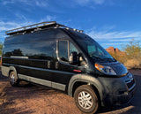 Black camper van RV rental with roof rack, on rugged terrain under blue sky, perfect for outdoor exploration.