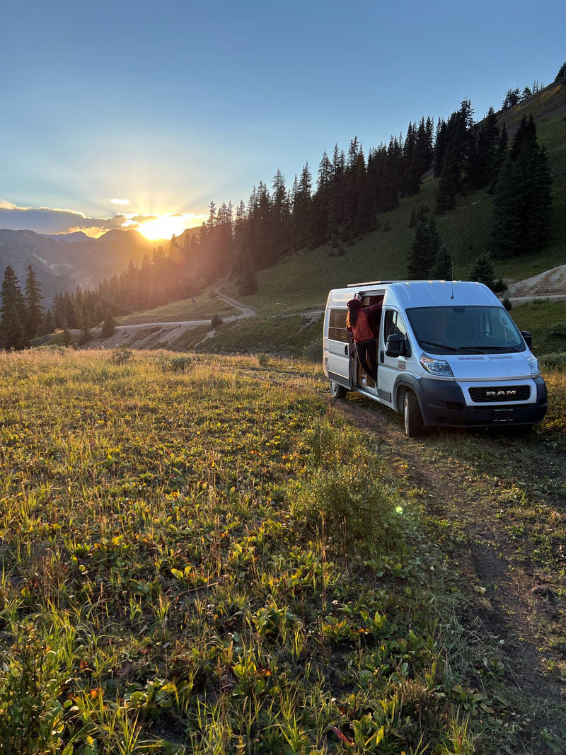 RV rental campervan adventure: A person exits the van in a sunlit mountain meadow with pines at sunset.|Scenic RV rental adventure: a man stands in a silver camper van — photo 1