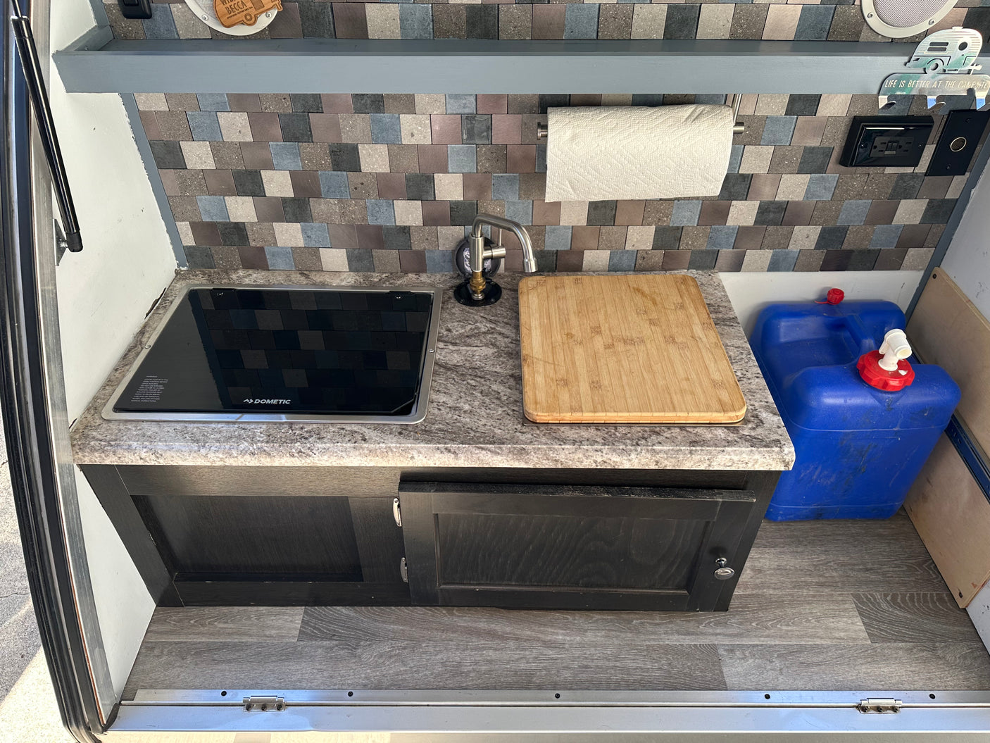 RV rental: compact kitchen with Dometic cooktop, sink, tiled backsplash, and blue water tanks for water supply.