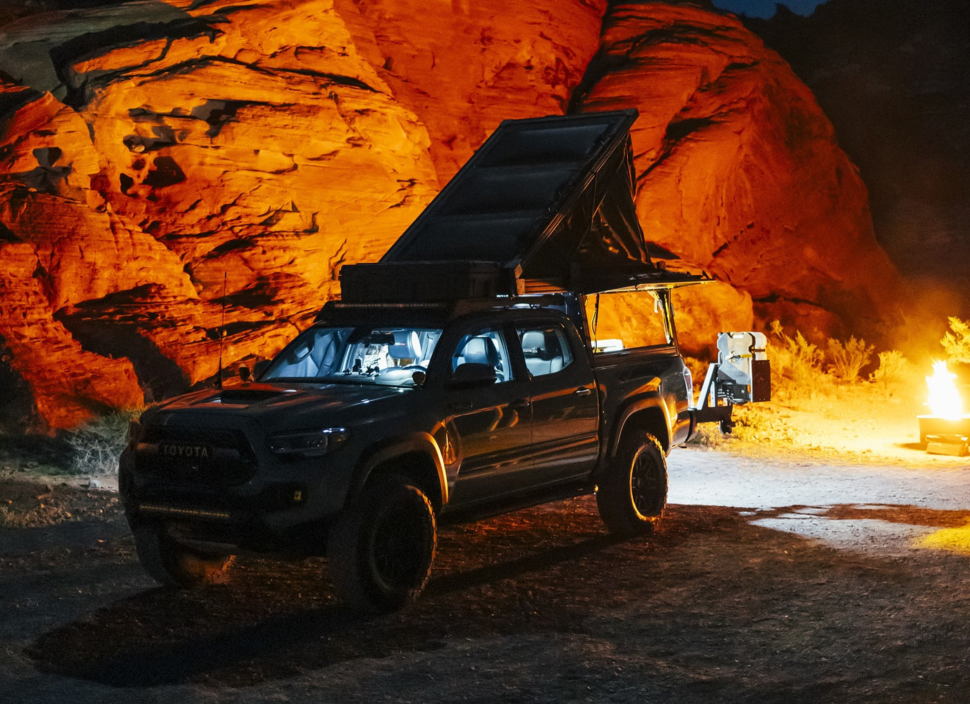 Adventure-ready RV rental: a Tacoma with a rooftop tent, lit by campfire glow & orange rocks at night.