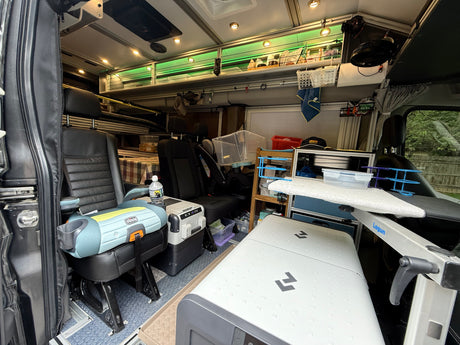 This RV rental interior features a Chicco car seat, portable fridge, Lagun table, and organized overhead storage with green LEDs.