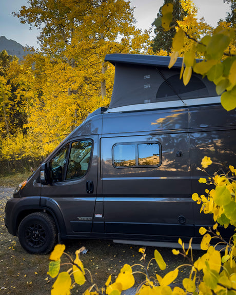 Grey RV rental with pop-top tent immersed in vibrant golden aspen trees, reflecting mountains & sky, perfect for fall adventures.