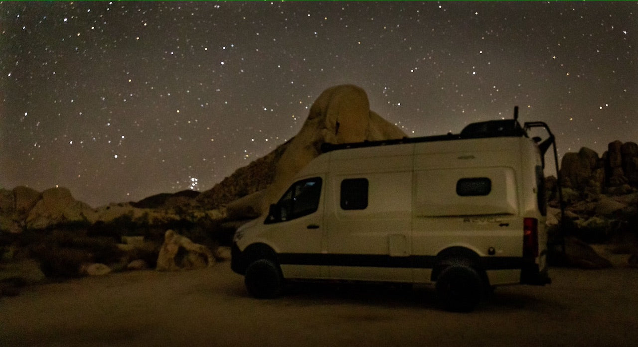 A white RV rental camper van is parked beneath a magnificent starry night sky, surrounded by rugged desert rock formations.