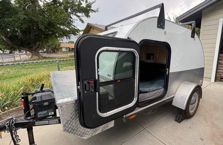 Teardrop camper, ideal for an rv rental. Door open showing cozy bed, front storage box, & battery. Ready for travel!