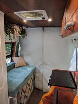 A cozy rv rental interior features a teal cushioned bench, wood-paneled ceiling with a fan, and a compact kitchen setup.