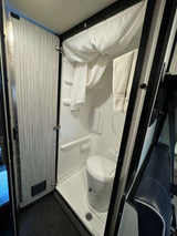 Compact RV rental bathroom: white shower basin, toilet, and a wood-patterned folding door optimize space for travelers.