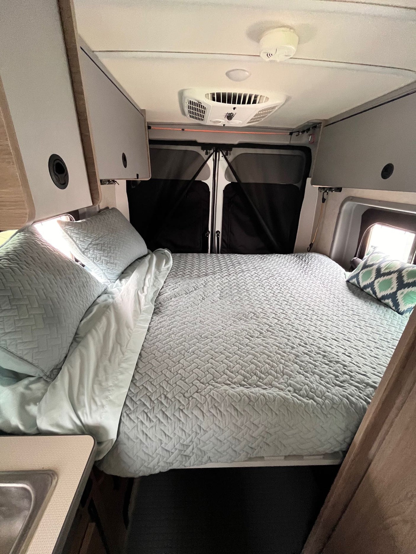 Inside a comfortable RV rental: a large bed with light blue bedding, overhead cabinets, and rear door covers.