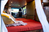 Inside this cozy RV rental: a custom van conversion features a comfy bed with orange cushions and wood accents.