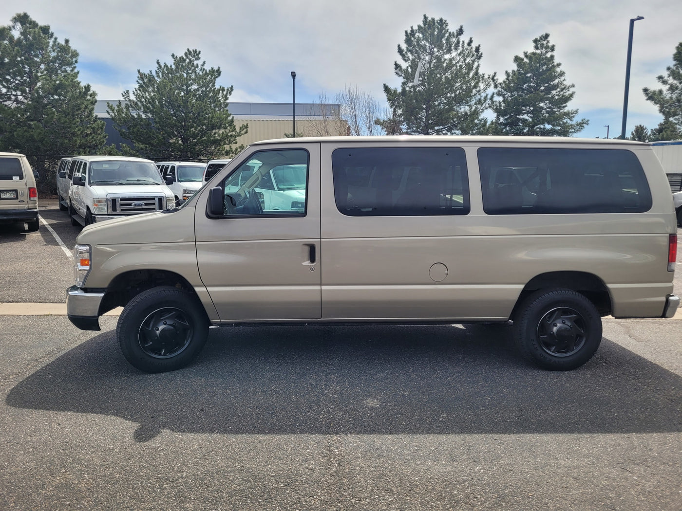 A beige passenger van, often found in RV rental fleets, parked in a lot with other vans and trees. — photo 9