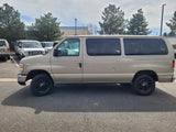 A beige passenger van, often found in RV rental fleets, parked in a lot with other vans and trees.