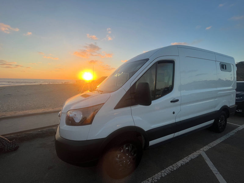  with a warm campfire in the foreground and a picnic table in the mid-ground.|White Ford Transit RV rental at a waterfront campsite (site 55) with city lights across the bay. Windshield frosted.|An RV rental van overlooks the bay — photo 12