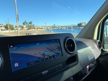 RV rental dashboard with navigation showing a coastal route. Outside, a sunny beach and boats in a bay under blue skies.