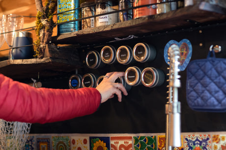 Person's hand reaches for a spice jar on a magnetic rack, showcasing compact kitchen organization in an RV rental. Cooking essentials.